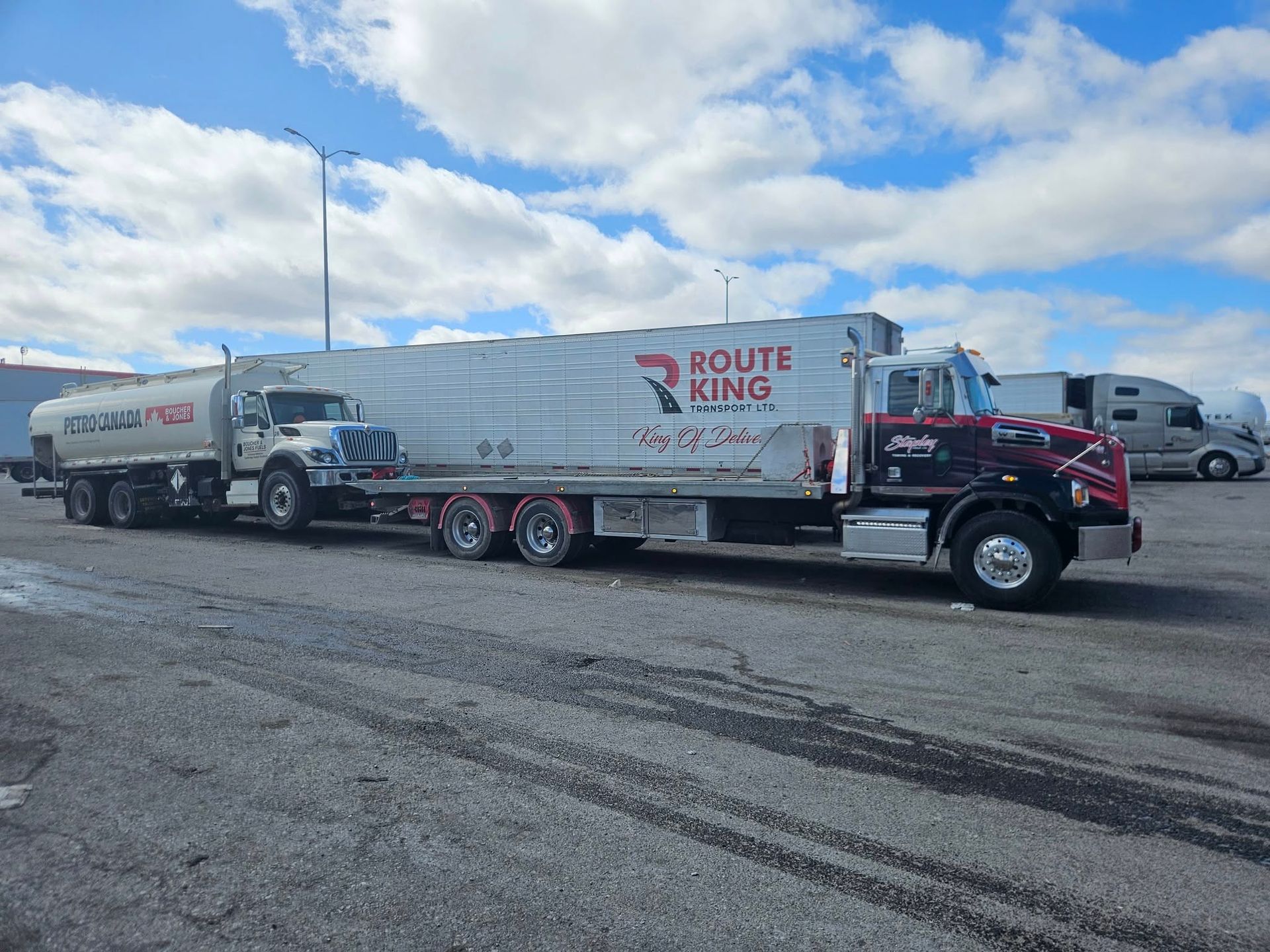 A semi-truck with a Route King trailer and a tanker truck in a parking lot under a cloudy sky.