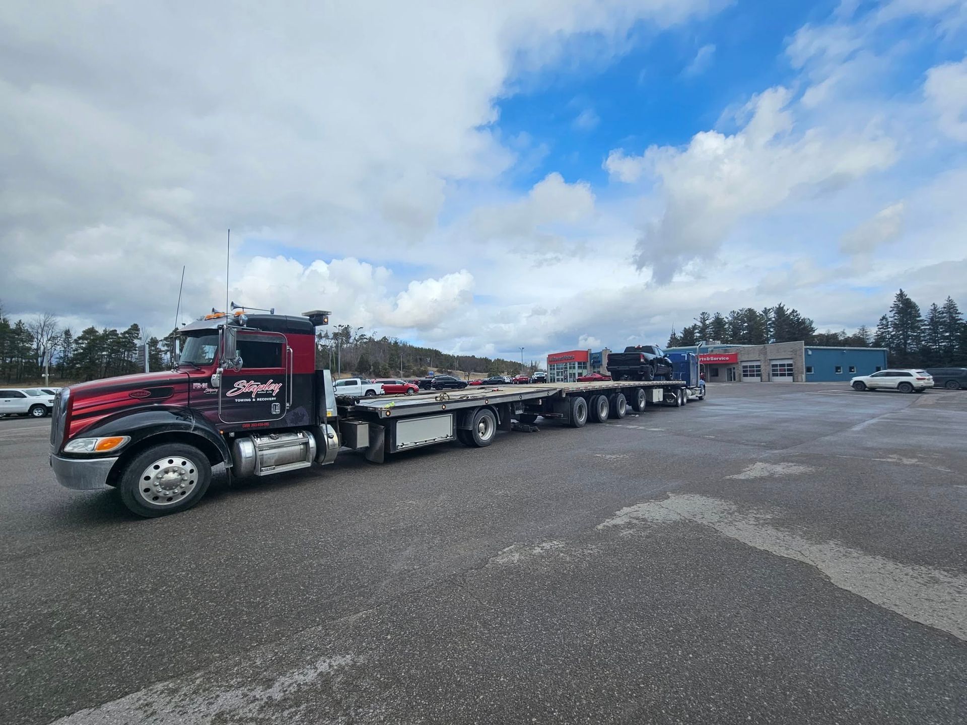 Black and red semi-truck with a long flatbed trailer parked in a lot under a cloudy sky.
