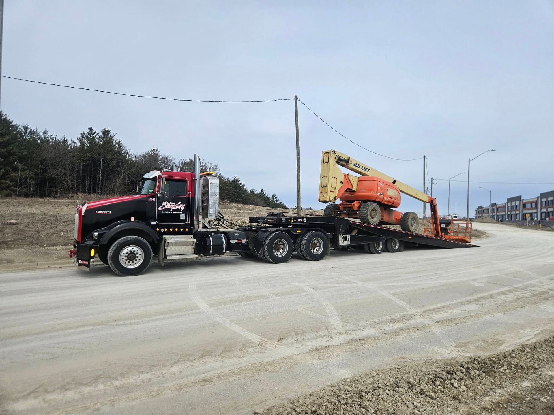 A red and black semi-truck hauling an orange lift on a lowboy trailer on a dirt road.