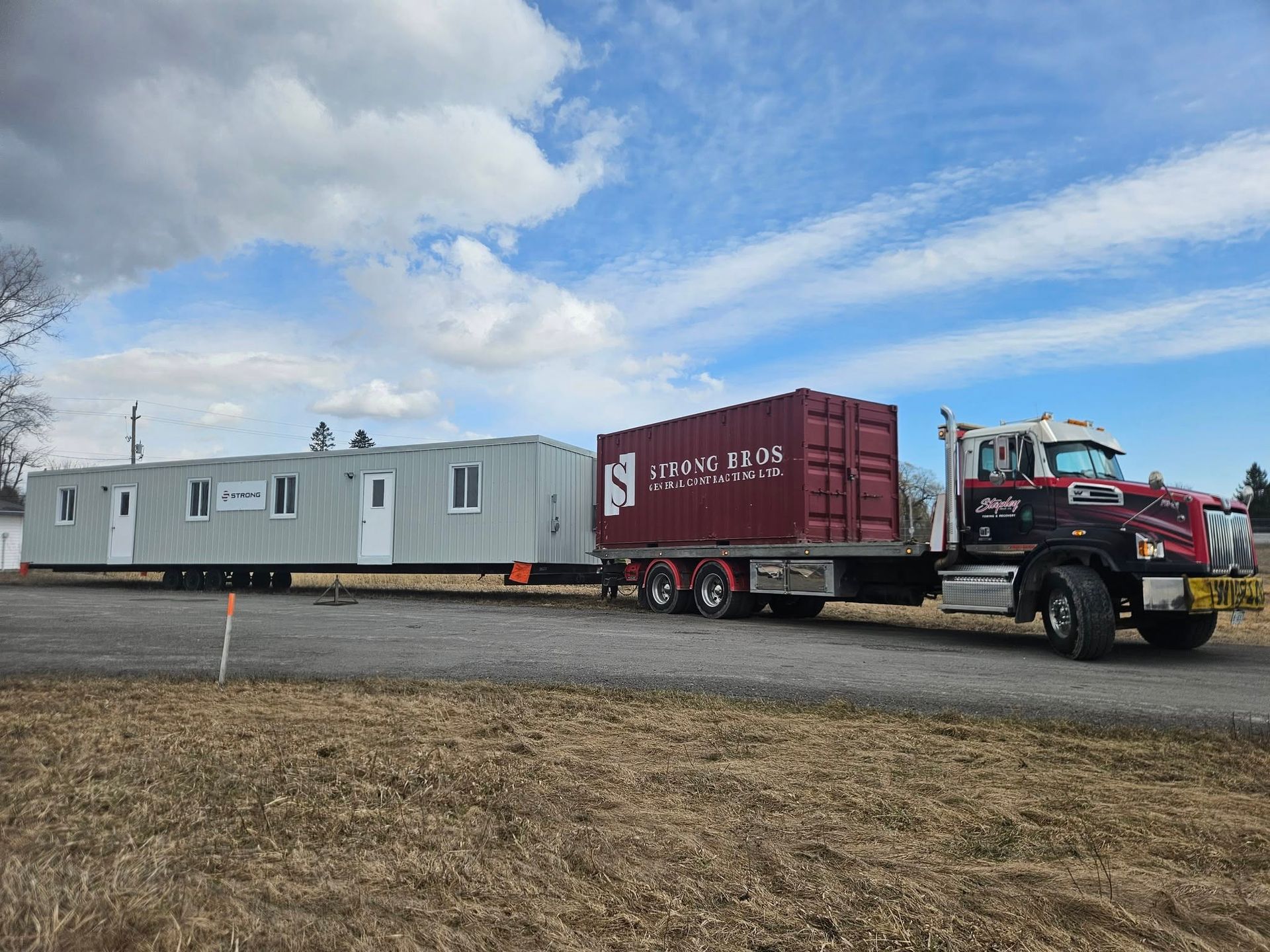 Semi-truck hauling a long, modular building and a cargo container on a trailer, outdoors under a cloudy sky.
