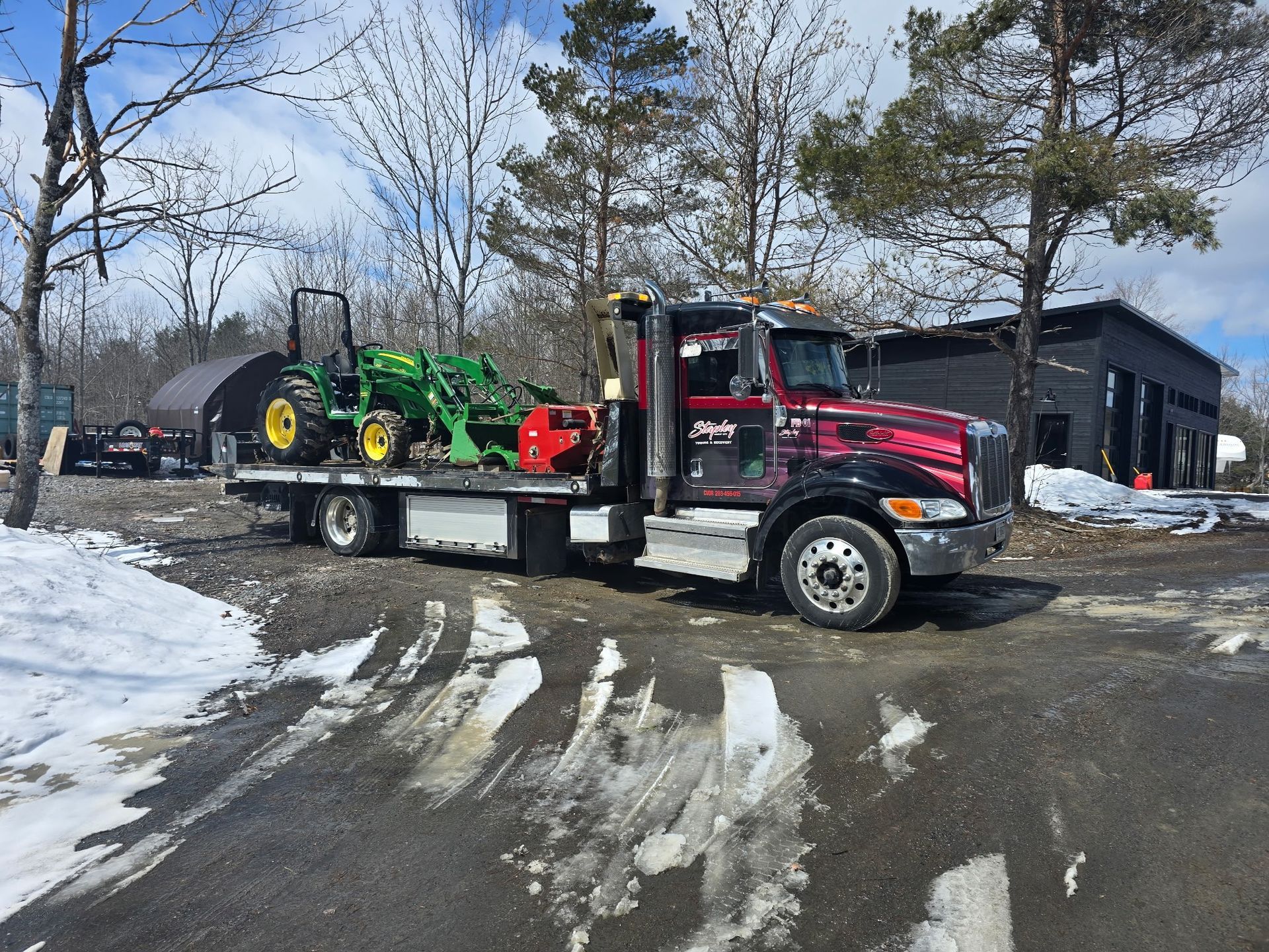 Tow truck hauling a green and yellow tractor with a snow-covered parking lot and dark building in the background.