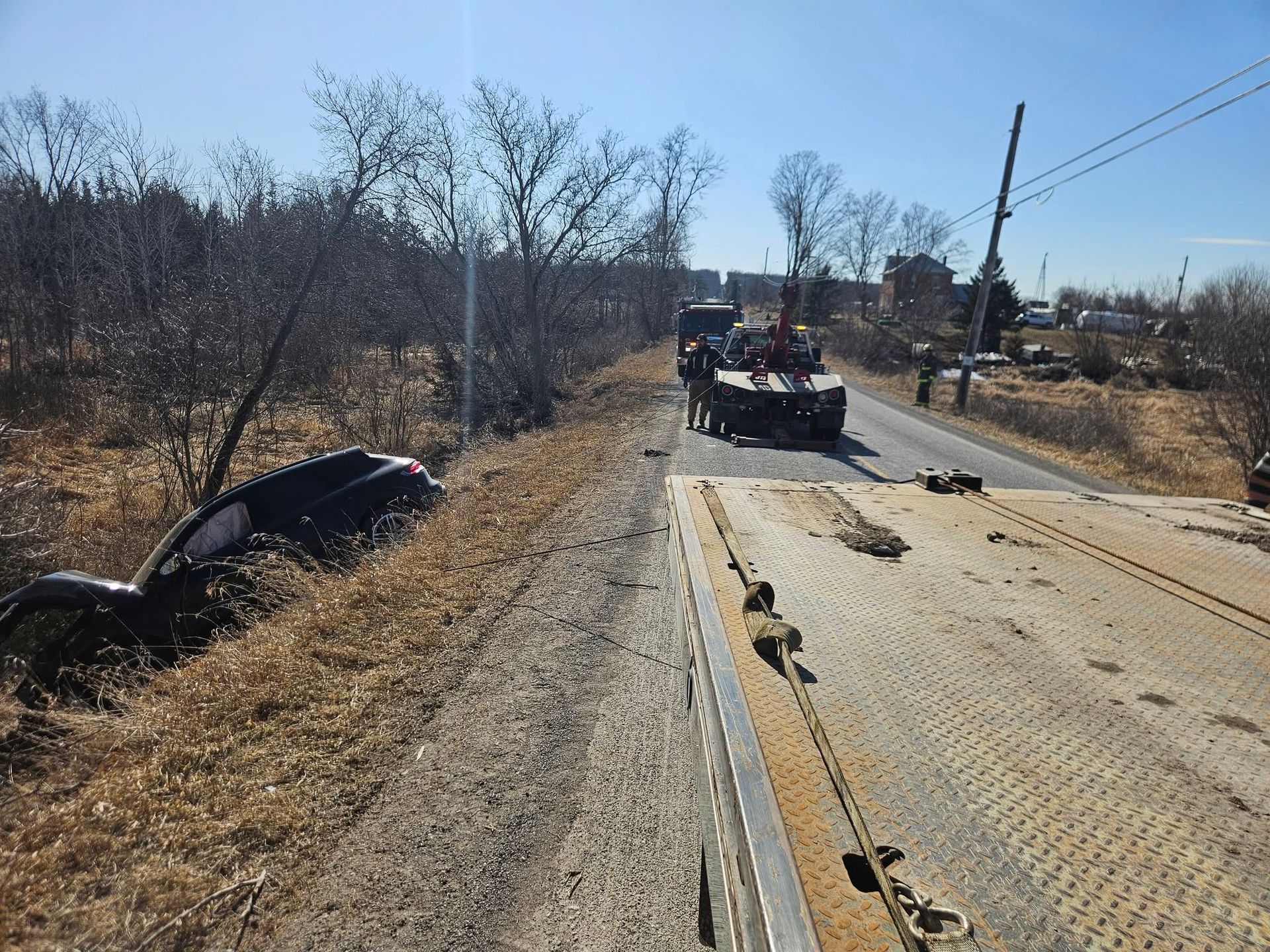 Car in ditch being towed away by a truck on a roadside with dry grass and trees.
