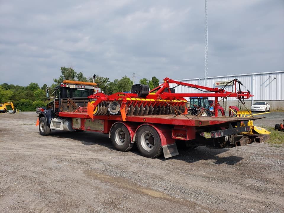 Red flatbed truck hauling farm equipment on a gravel lot under a cloudy sky.
