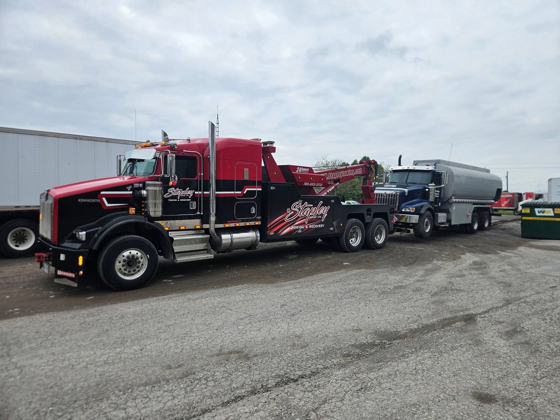 A large black and red tow truck towing a tanker truck on a gravel lot under a cloudy sky.