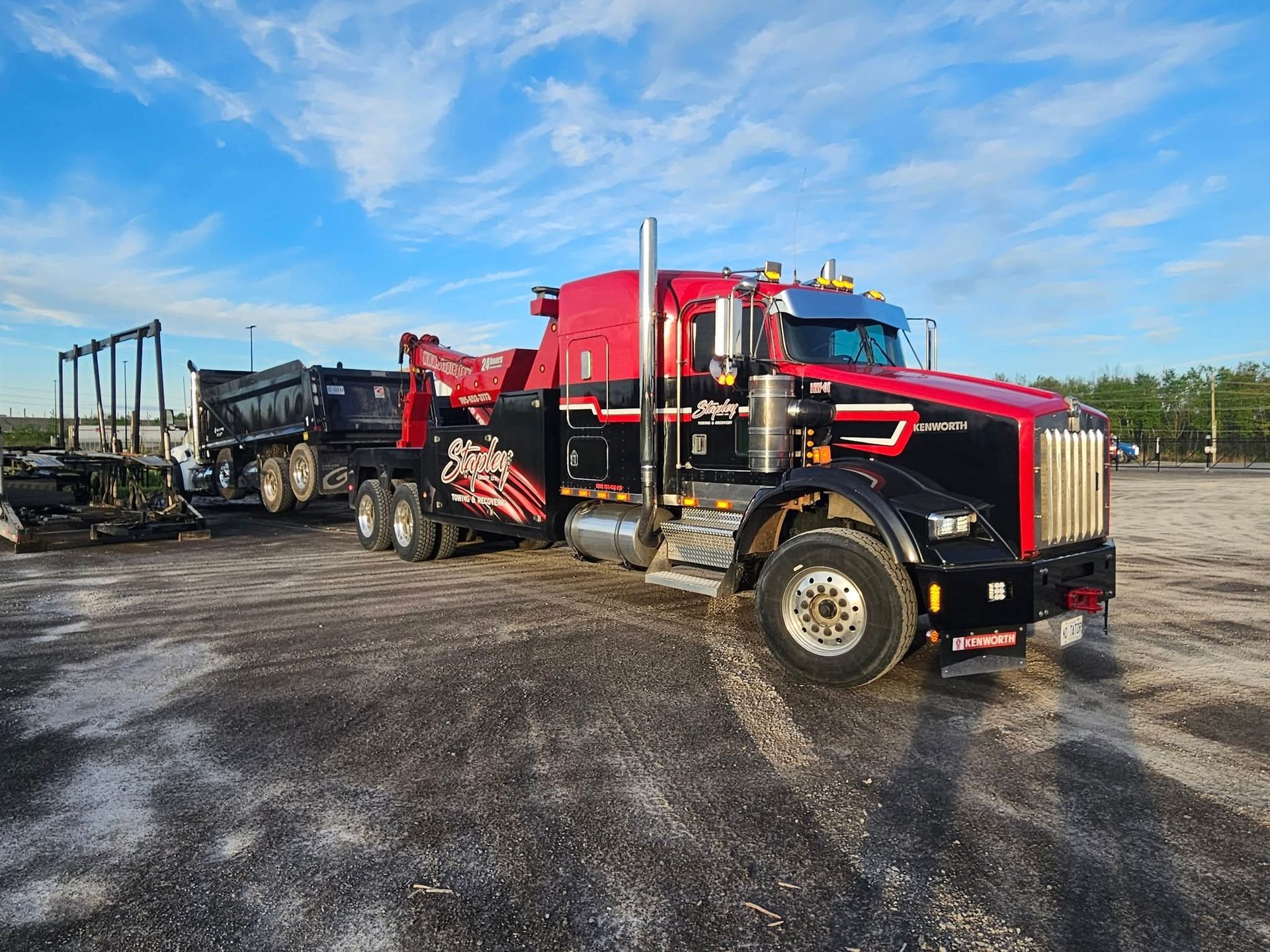 Black and red tow truck with a dump trailer on a gravel lot under a blue sky.