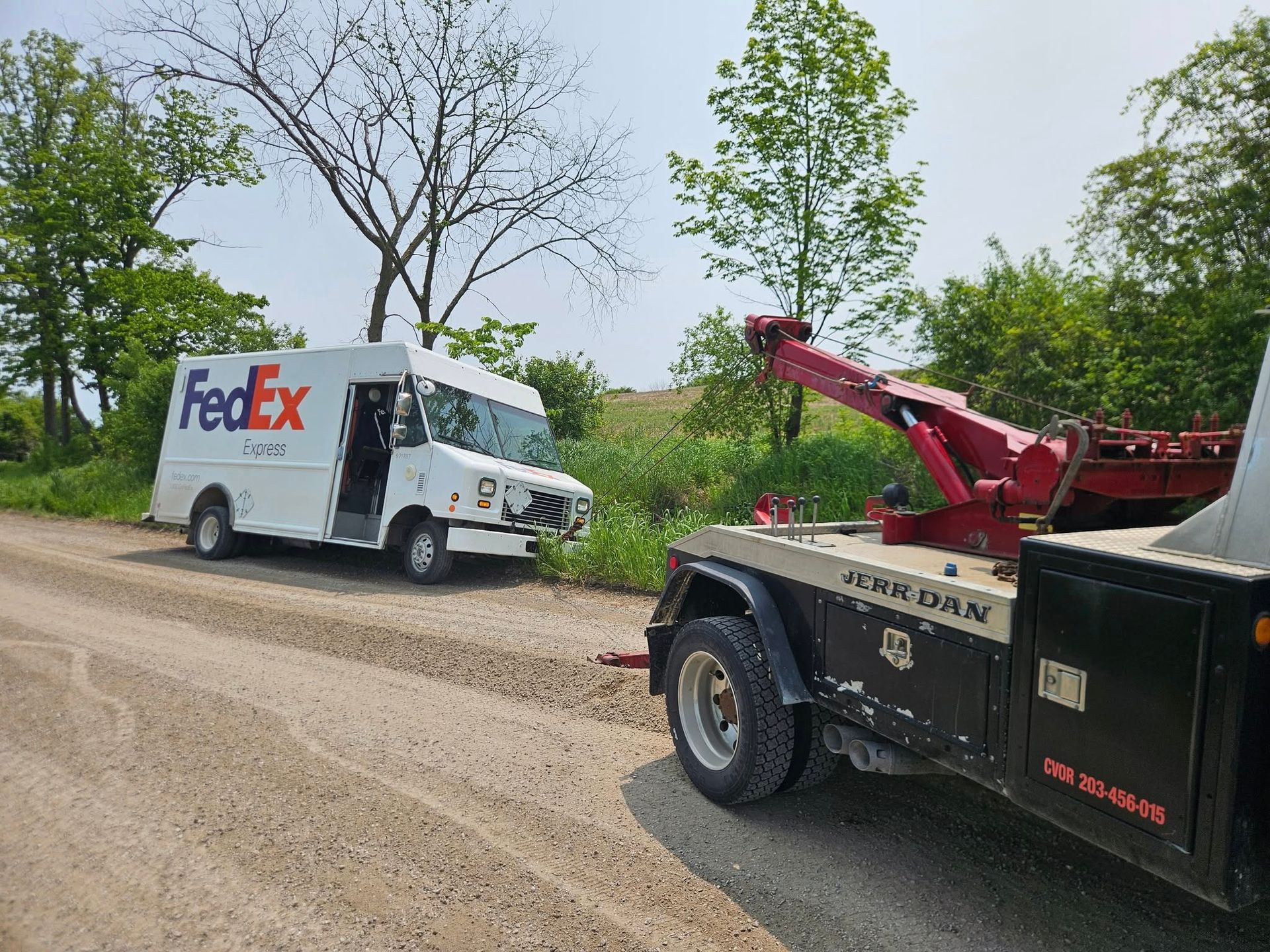 FedEx delivery truck being towed from a roadside ditch by a tow truck on a dirt road.