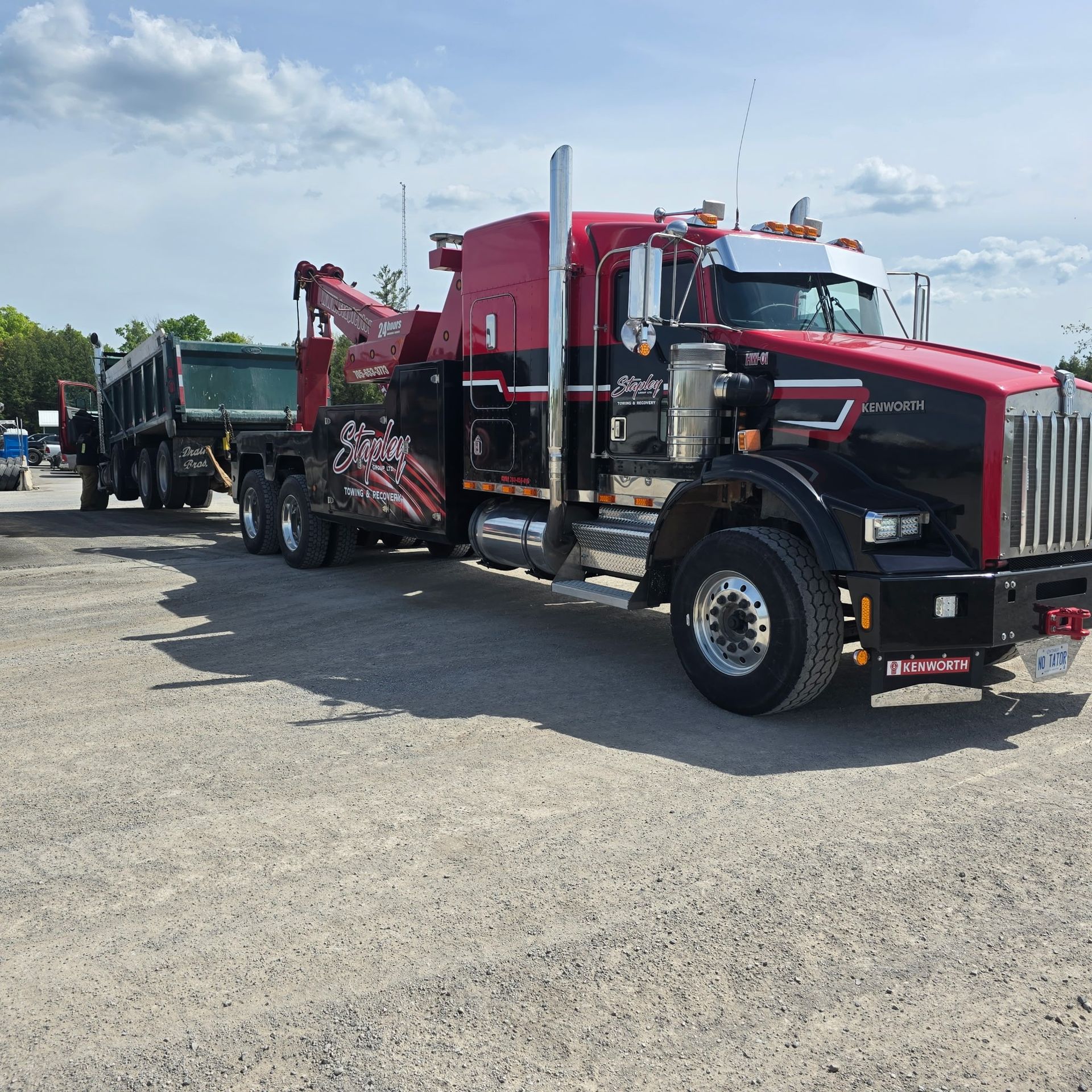 Black and red tow truck with attached dump trailer on gravel.