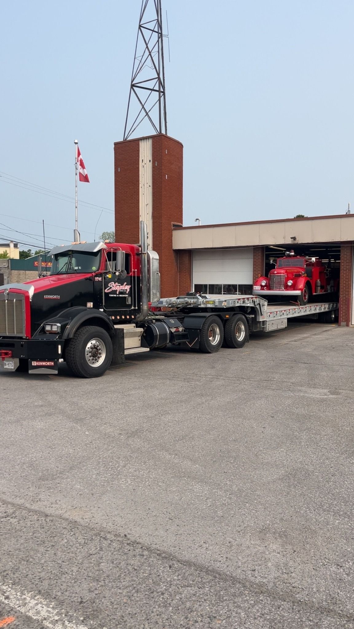 Red semi-truck with flatbed pulling a vintage fire truck out of a brick fire station.