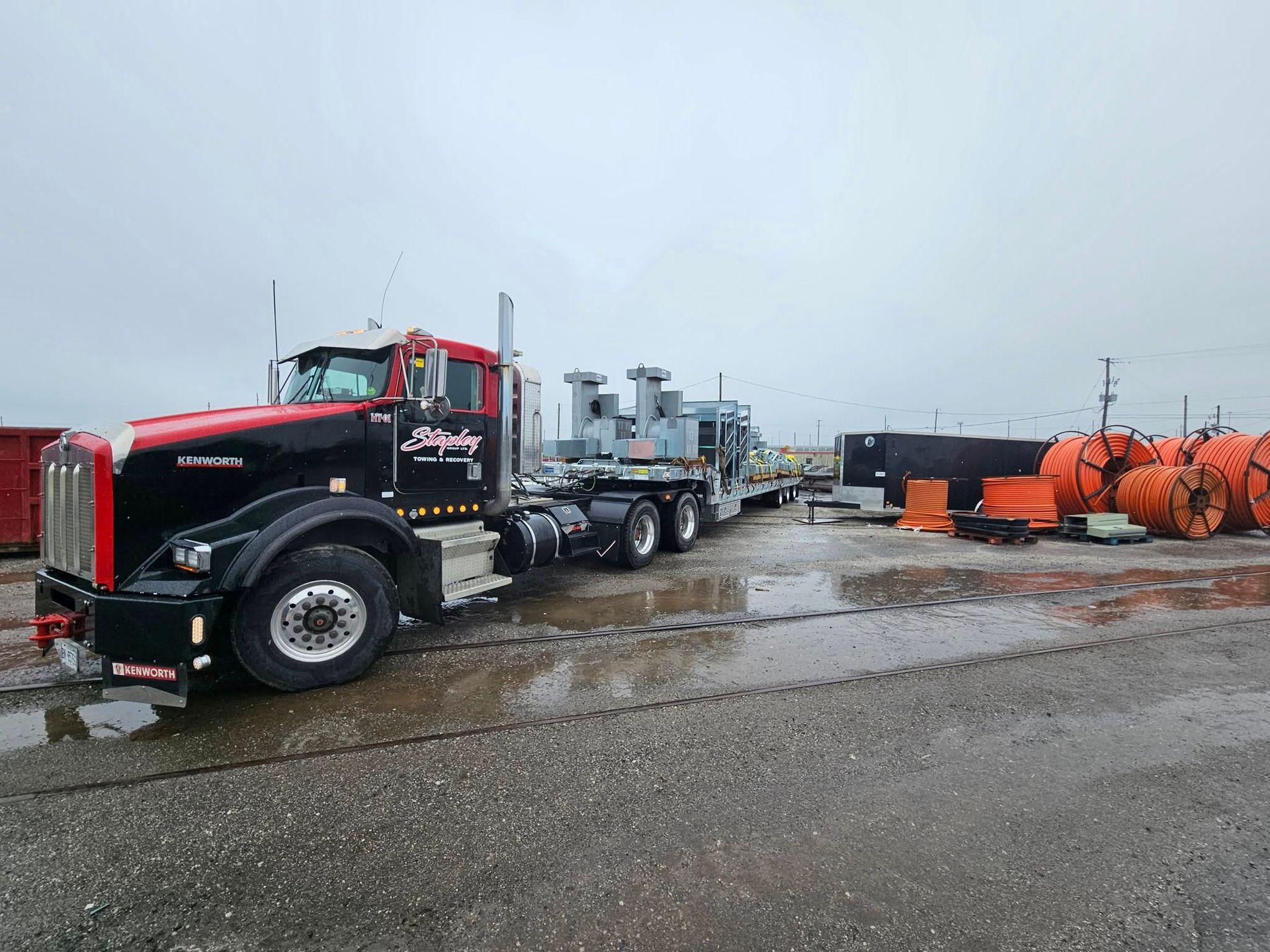 Black semi-truck hauling large industrial equipment on a flatbed trailer, parked on a wet, gray surface.