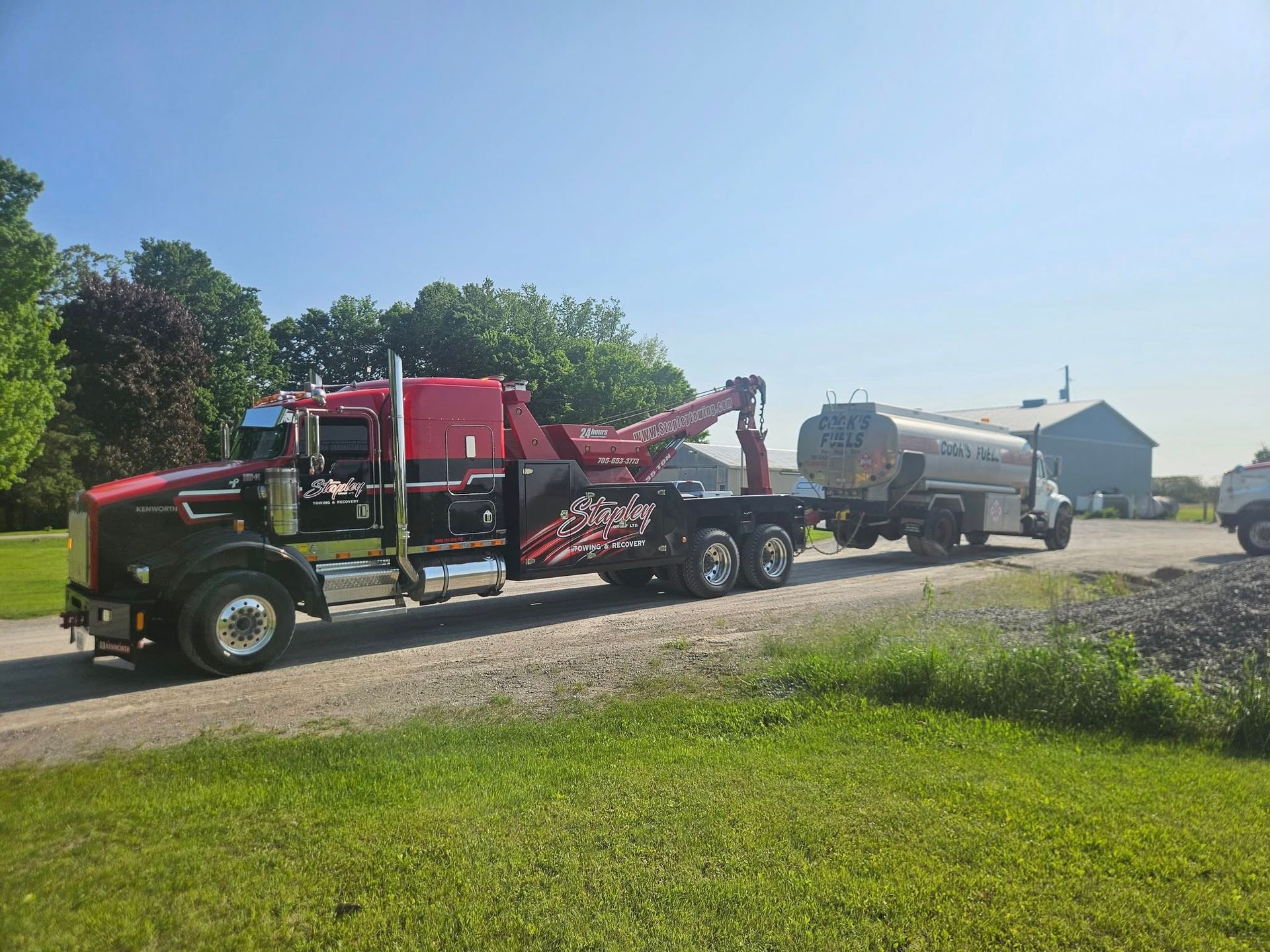 Red tow truck pulling a tanker truck on a gravel road, sunny day.