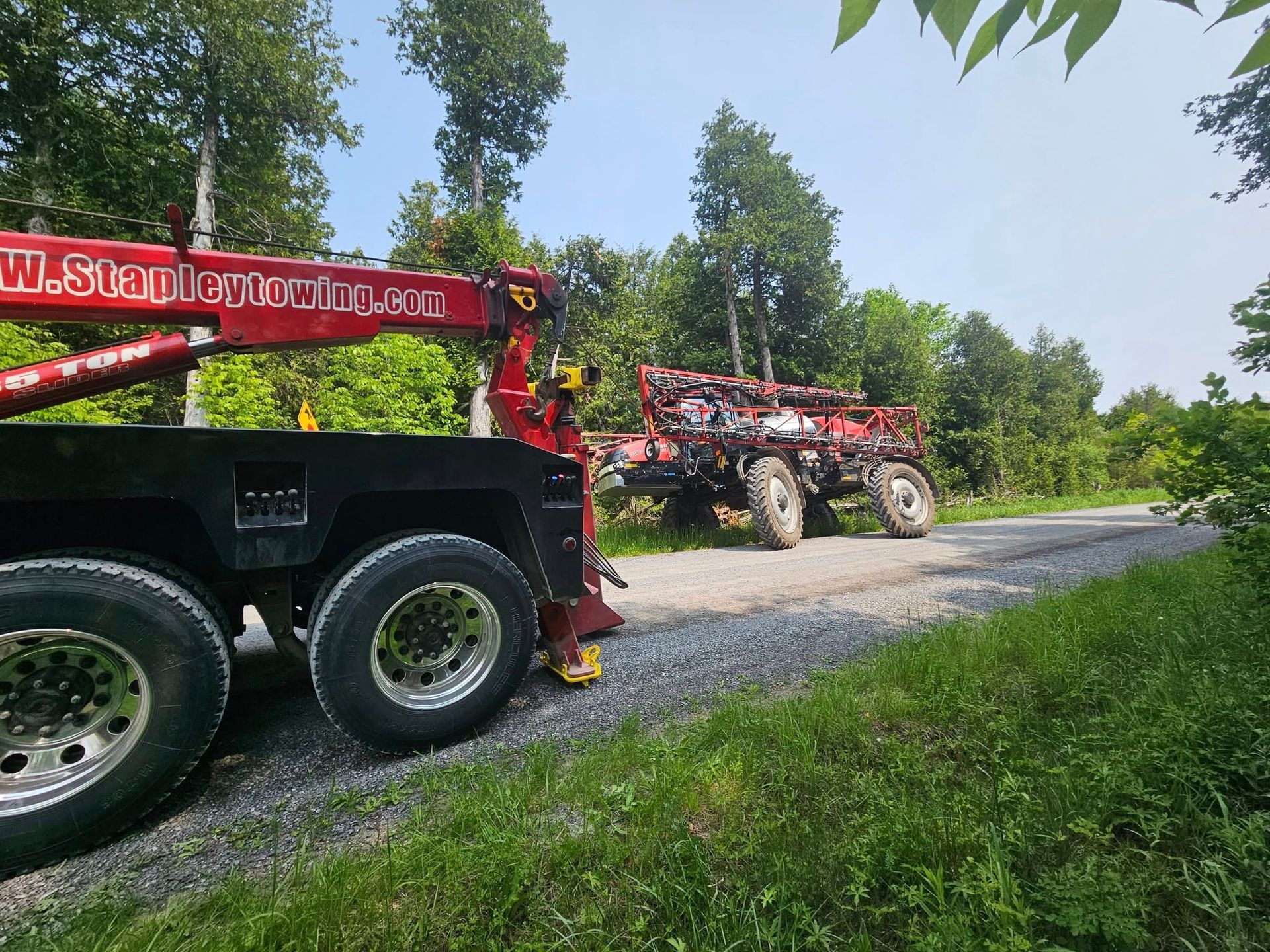 Red tow truck towing a damaged, red trailer on a gravel road, trees in the background under a blue sky.