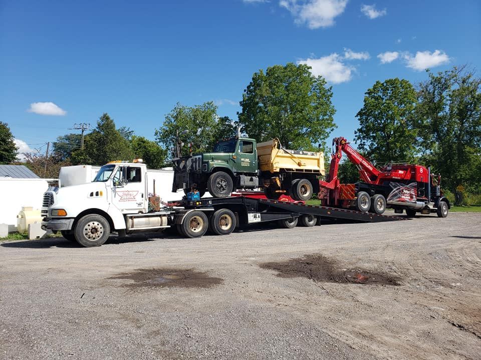 A white flatbed truck hauling a green dump truck and a red excavator under a blue sky.
