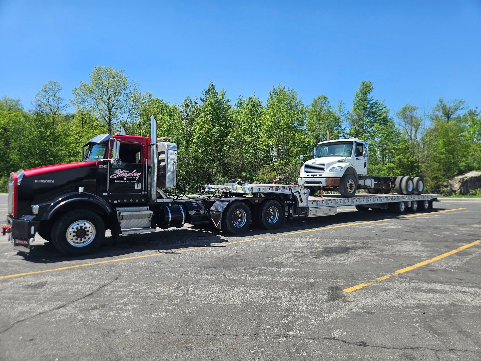 Black and red semi-truck towing a white truck on a flatbed trailer in a parking lot on a sunny day.