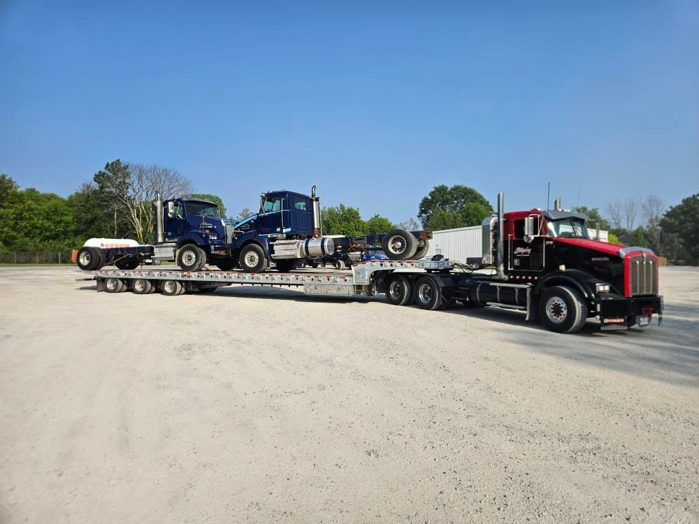 Semi-truck hauling two blue semi-truck cabs on a flatbed trailer in a gravel lot.