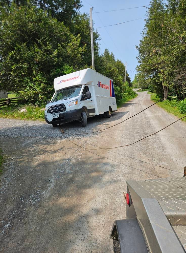 White delivery van being towed on a dirt road by a trailer. Trees line the sides; sunny day.