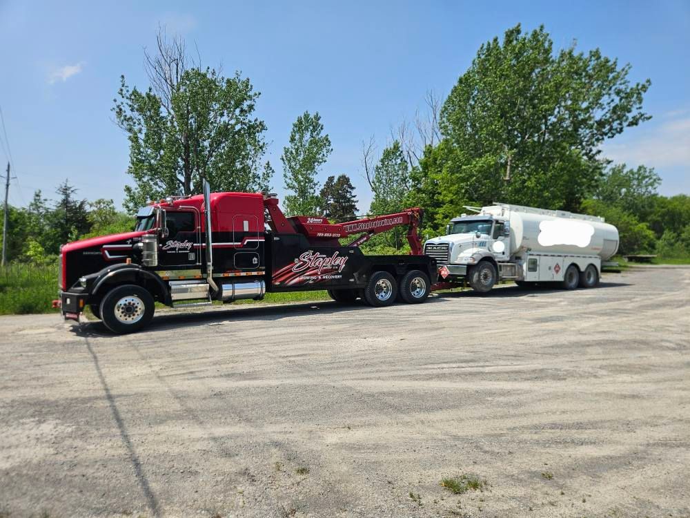 Tow truck hauling a white tanker truck on a sunny day. The tow truck is black and red.