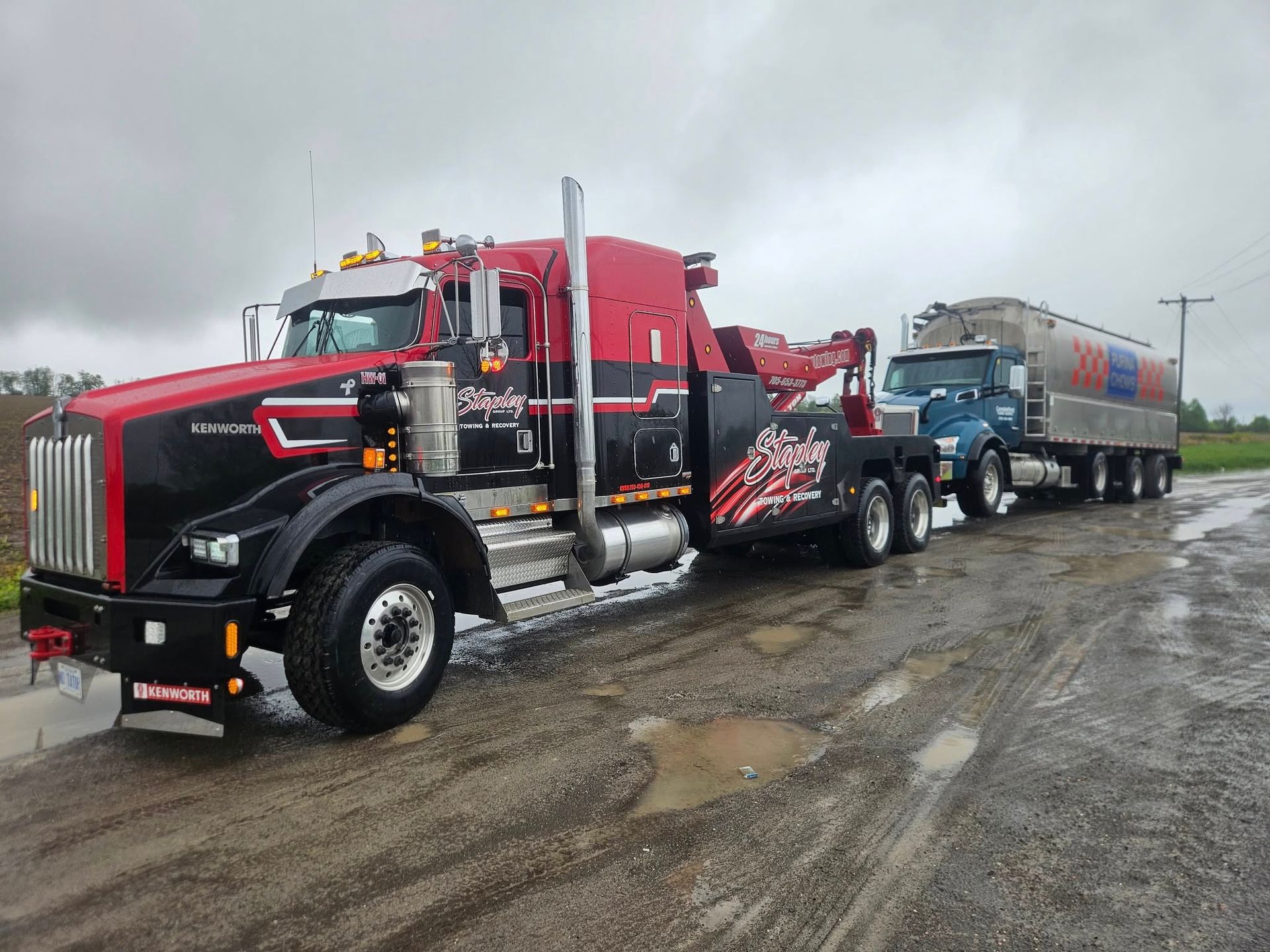 Black and red tow truck pulling a blue truck with a tanker trailer on a wet road.