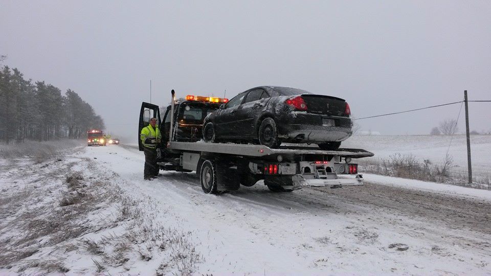 Tow truck loading a black car on a snowy, rural road. A person in a high-vis vest stands nearby.