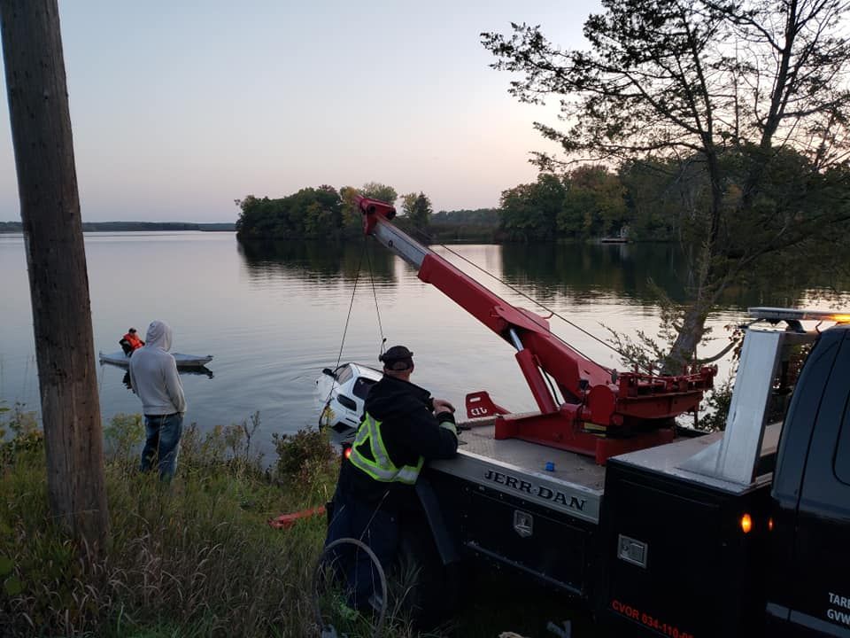 Tow truck lifting a car out of a lake. Two people watch from the shore. Red tow truck, water, trees.