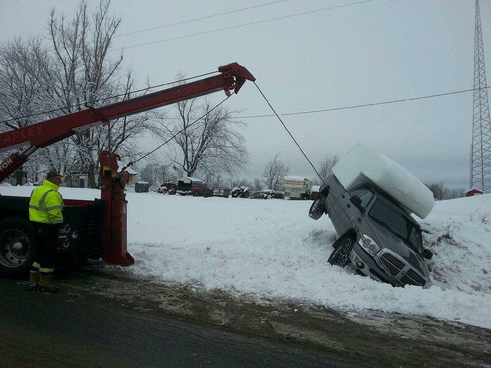 Tow truck lifting a gray SUV stuck in a snowbank near a road on a cloudy day.