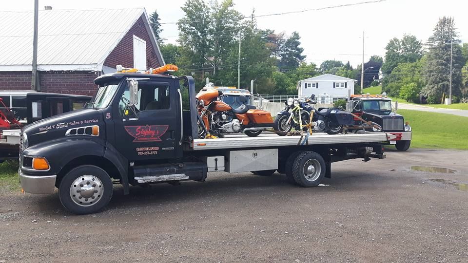 Black tow truck with two motorcycles on the flatbed, parked on gravel in front of buildings.