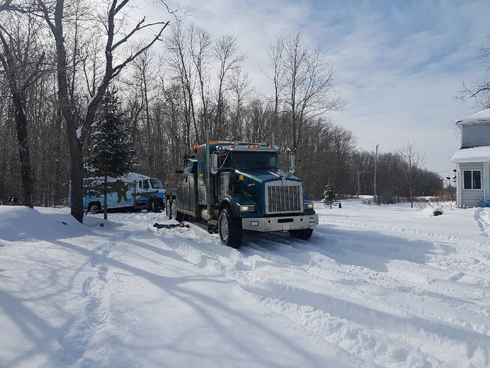 Tow truck driving in snow, near a house and trees.