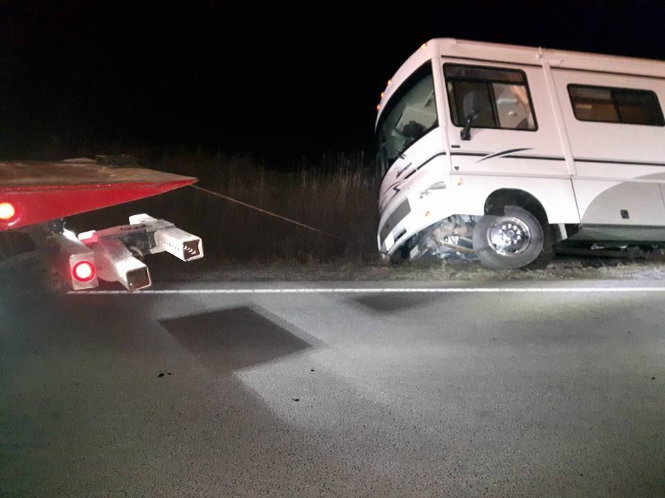 A white RV off the road with front-end damage, being towed by a red wrecker at night.