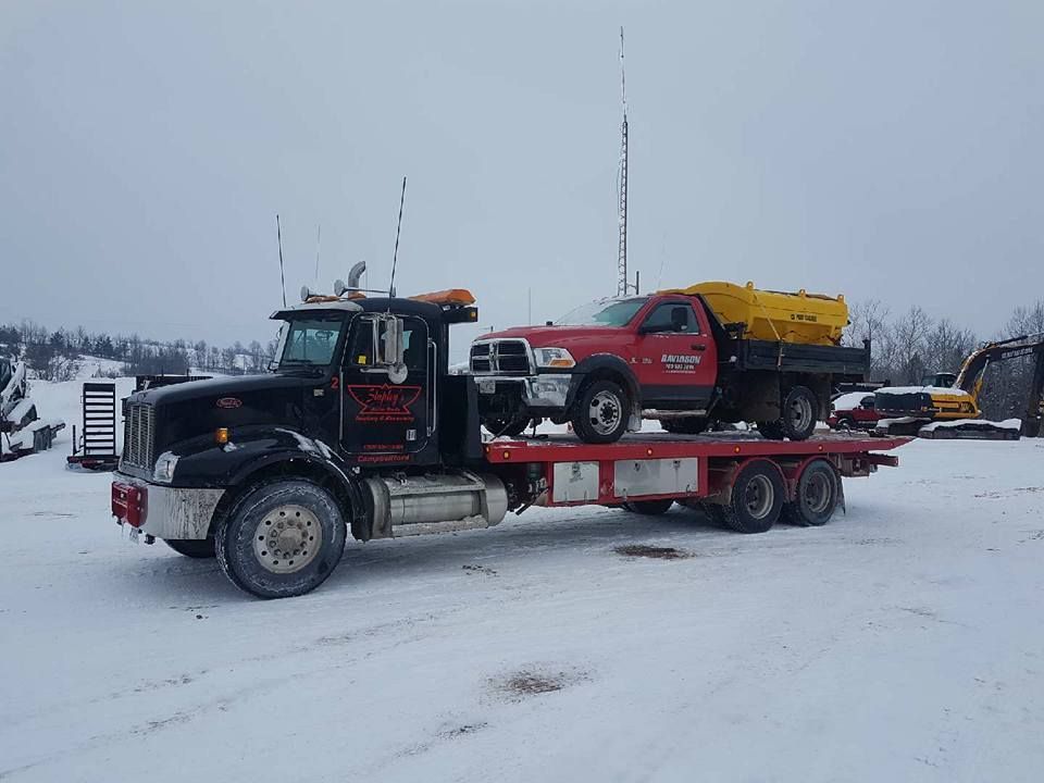 Black tow truck carrying a red pickup truck with a yellow salt spreader on a snowy day.