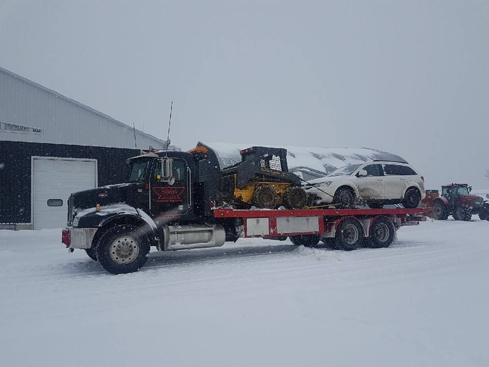 Flatbed tow truck carrying a skid steer loader and a white vehicle in a snowy setting.