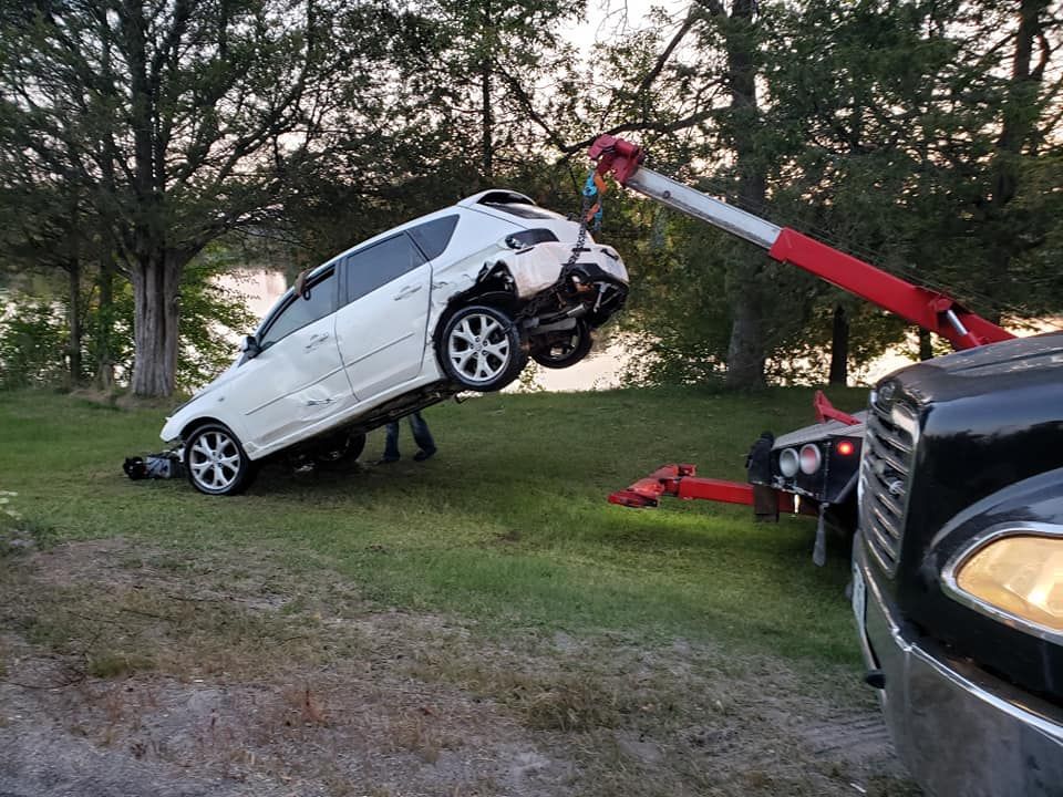A white SUV being lifted by a red tow truck on grassy land, near trees.