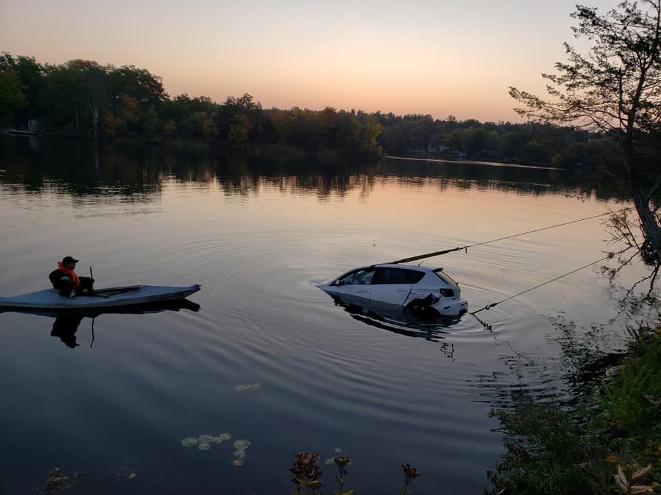 Car partially submerged in a lake, being pulled by ropes. A person in a kayak is nearby. Dusk.