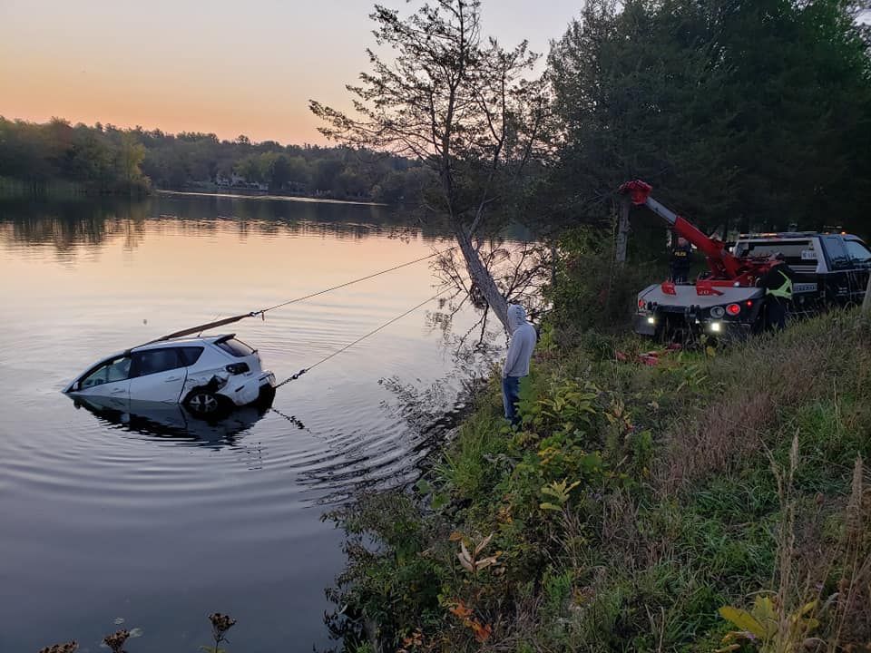 A white car submerged in a lake is being pulled out by a tow truck at dusk. A person stands nearby.
