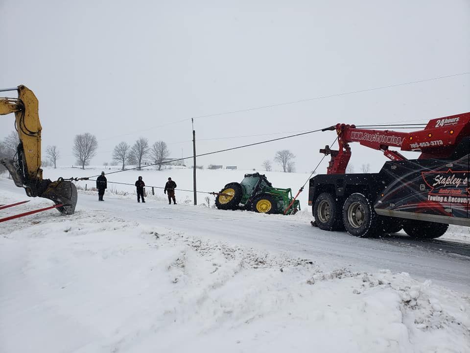 A tow truck is pulling a John Deere tractor out of a snow-covered ditch; several people watch nearby.