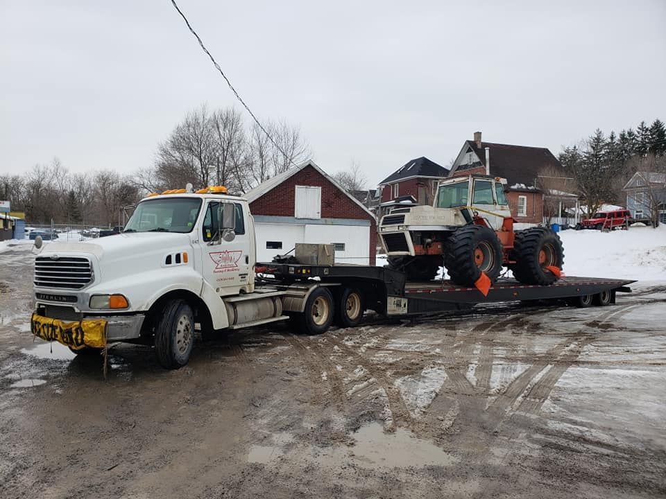 Tow truck hauling a white tractor on a trailer, outside in a snowy setting.