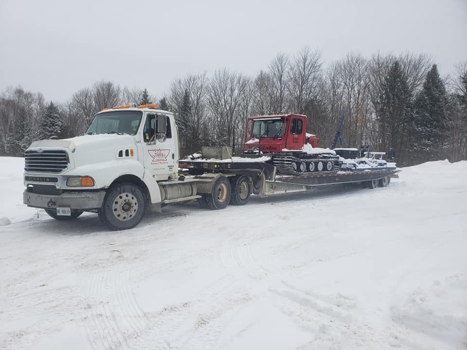 White truck towing a red tractor on a flatbed trailer in a snowy field.