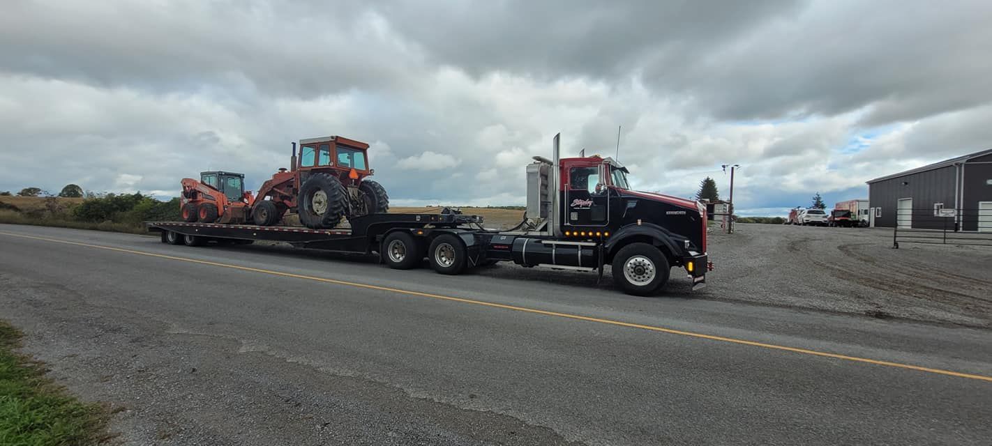 Black semi-truck hauling tractor and excavator on a lowboy trailer on a cloudy day.