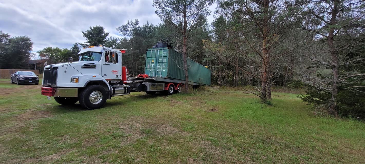 White truck hauling a large green shipping container through a grassy area near trees.