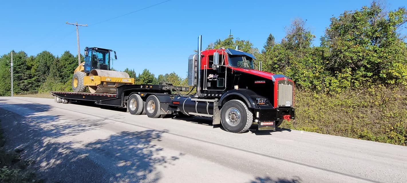 A black and red semi-truck hauling a yellow bulldozer on a flatbed trailer on a dirt road.