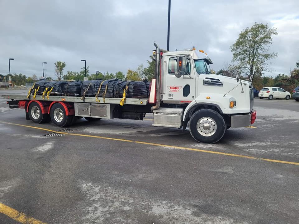 White flatbed truck loaded with black bags parked in a gray parking lot on an overcast day.