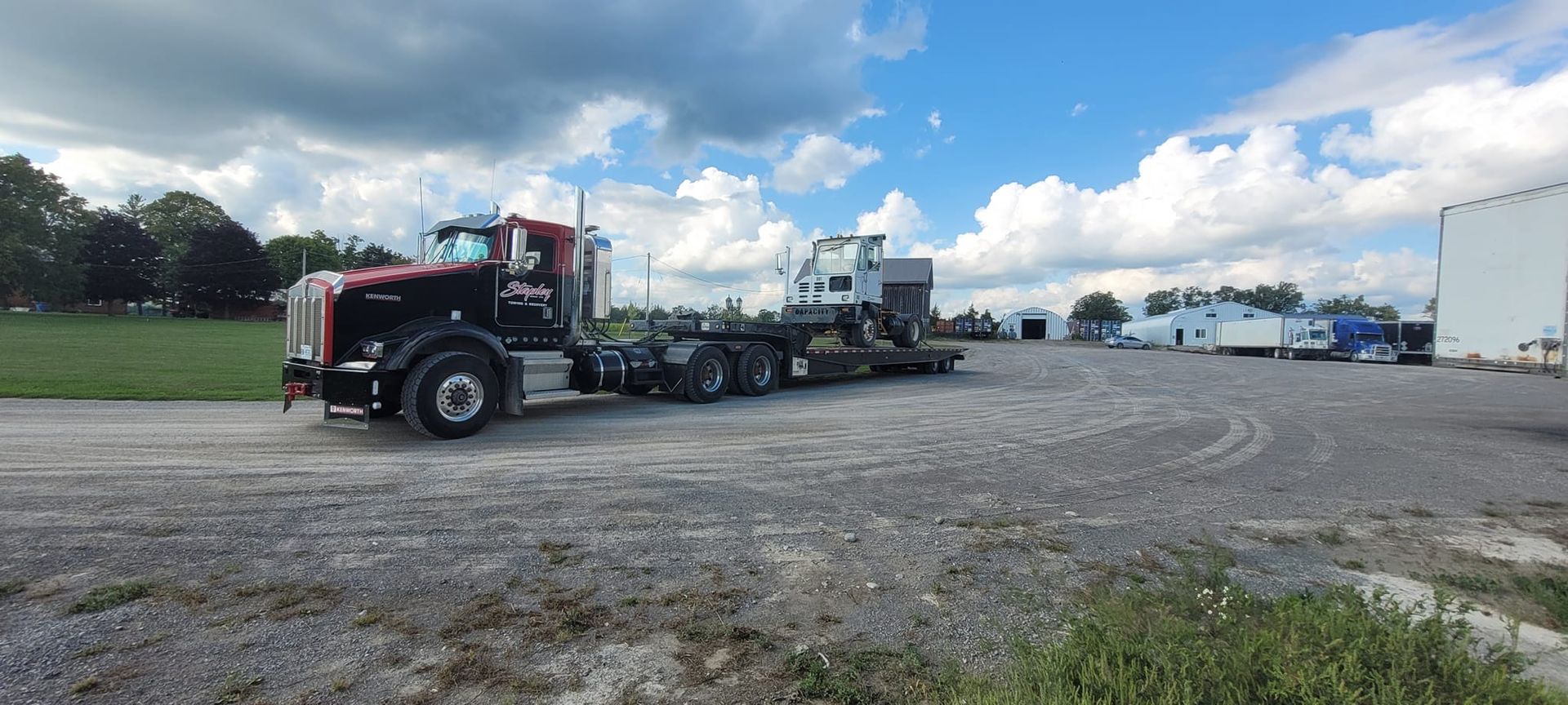 Black semi-truck hauling a white object on a gravel lot near a building under a cloudy sky.