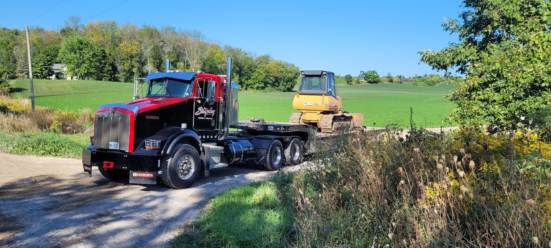 Semi-truck hauling a small excavator on a flatbed trailer on a dirt road, next to a field.