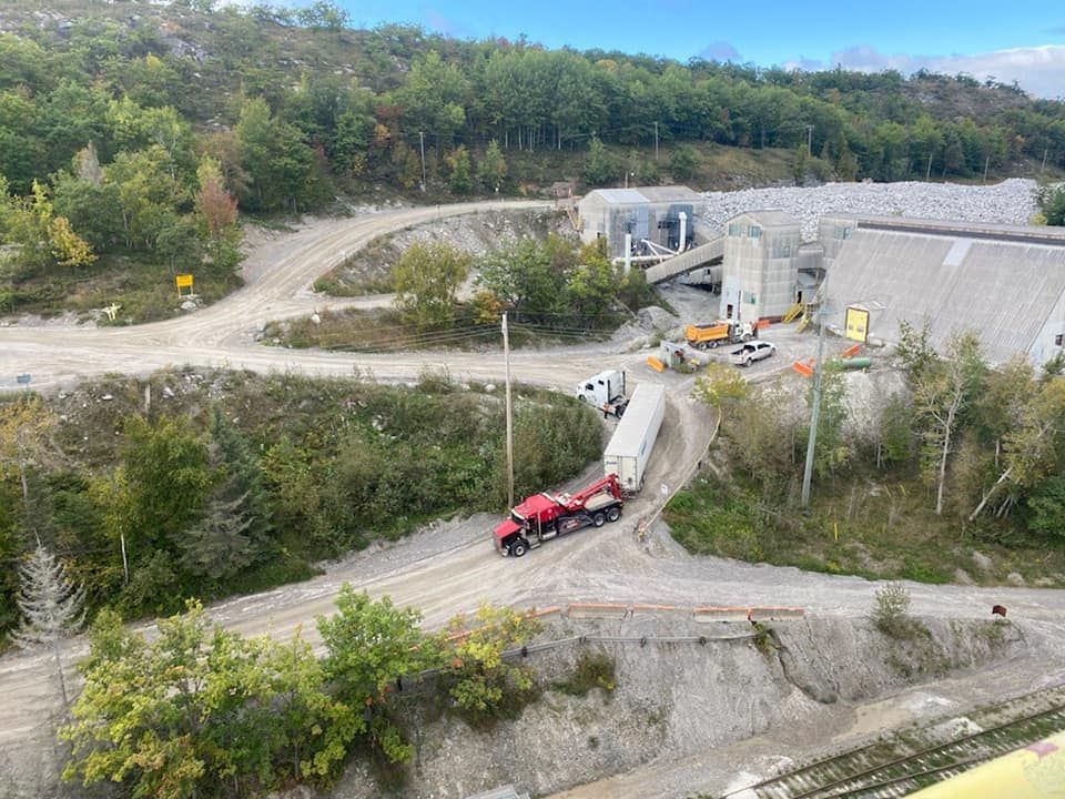 Red truck towing trailer on a gravel road near a quarry processing facility with buildings, trees, and a hillside.