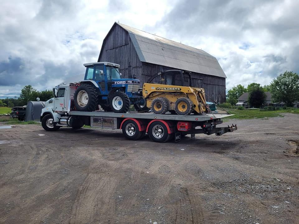 Flatbed truck carrying a blue tractor and yellow skid steer in front of a barn.