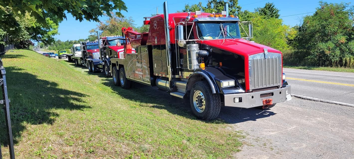 A red tow truck parked on the side of a road, with other vehicles behind it. Green grass and trees are present.