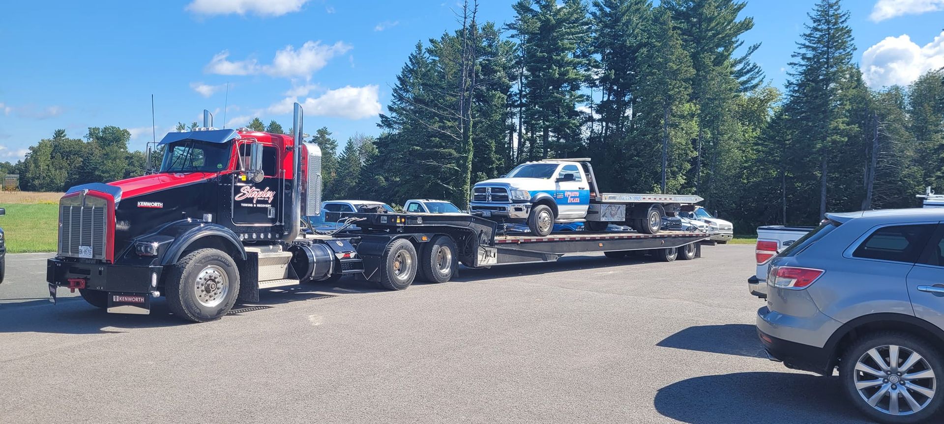 A black and red semi-truck with a car hauler trailer is parked in a lot. A white truck and a silver car are on the trailer.