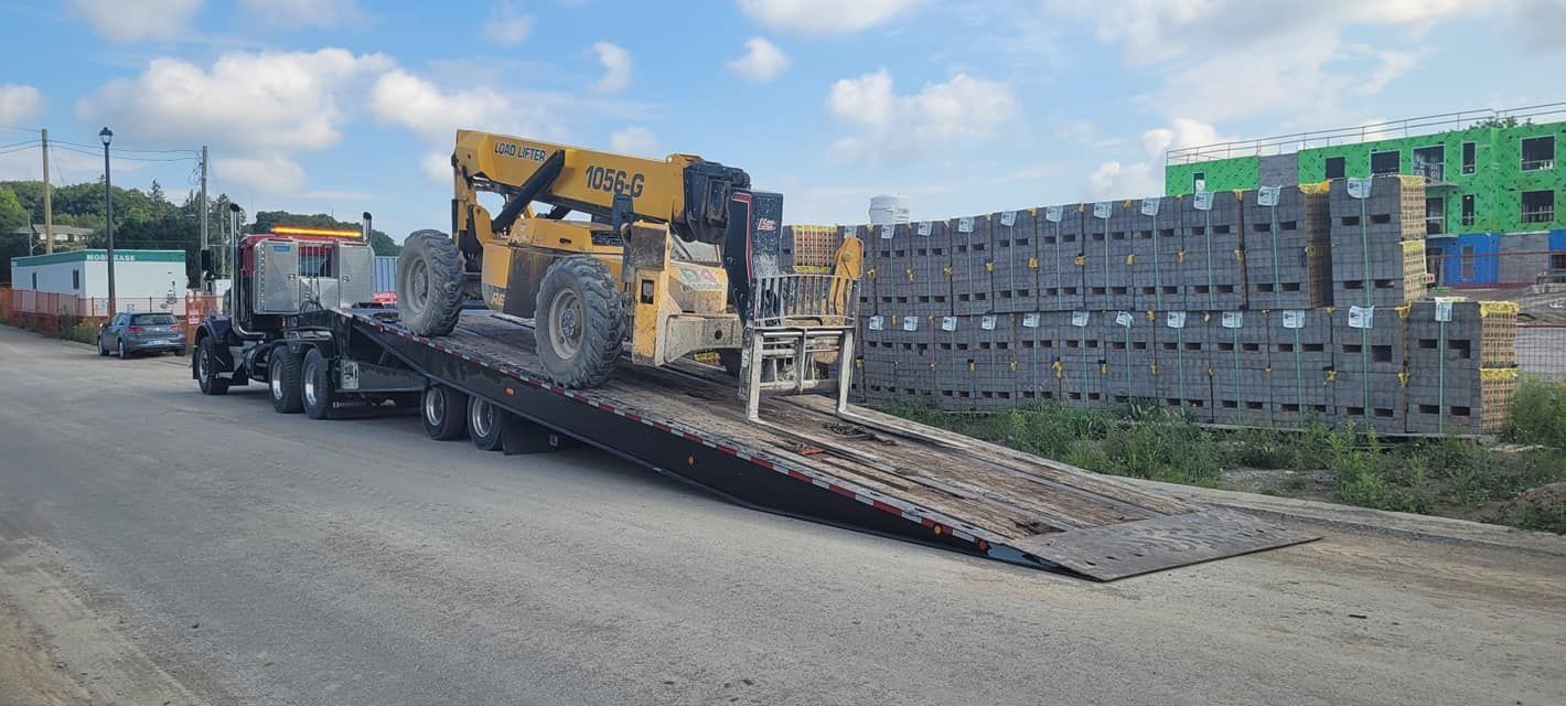 A semi-truck hauling a yellow construction vehicle on a flatbed trailer, next to stacked concrete blocks.