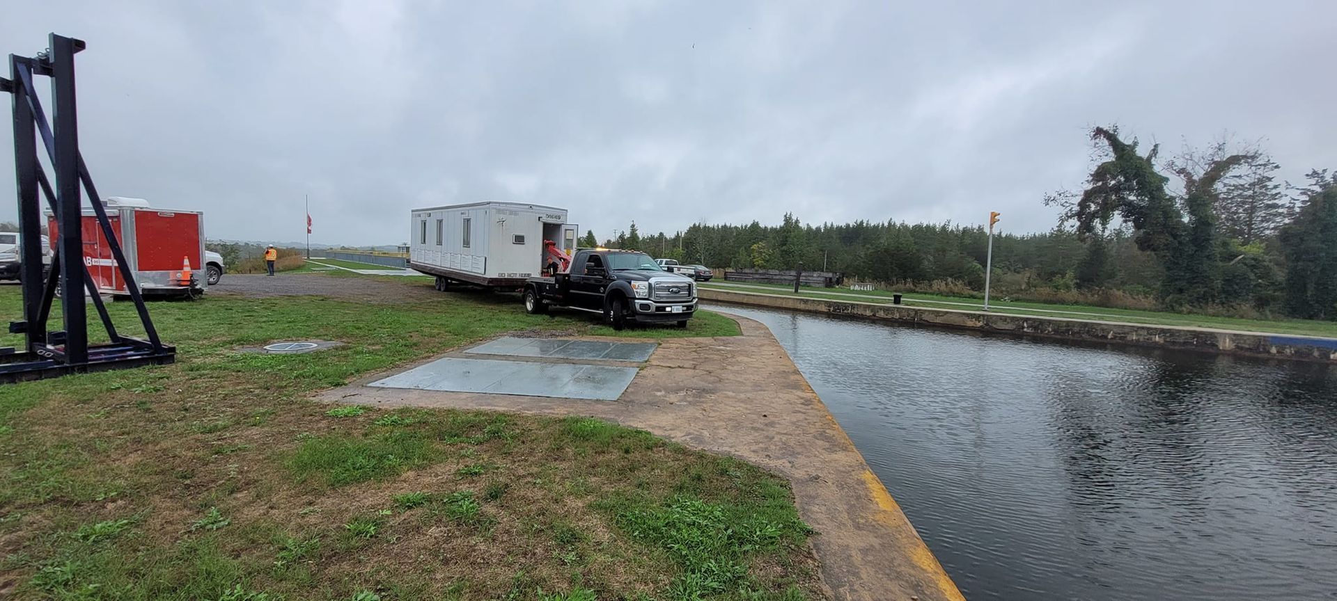 A truck towing a trailer on a grassy area next to a road, overcast sky.