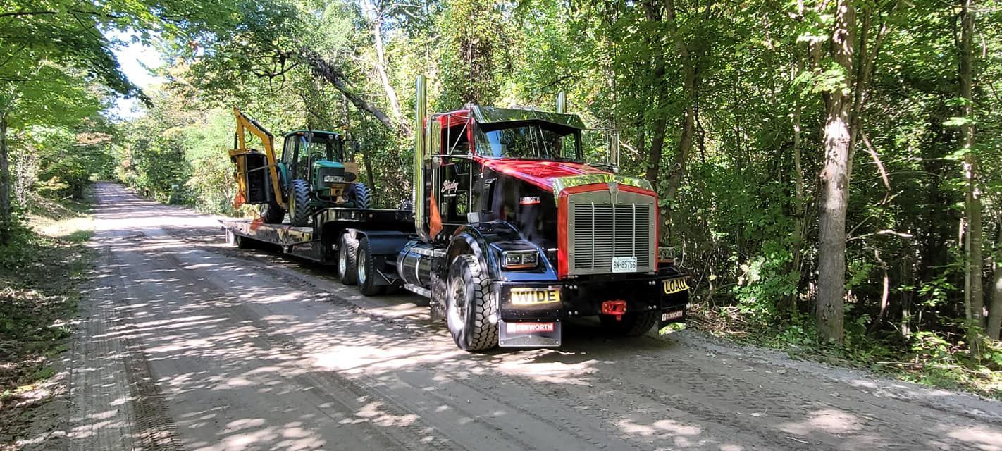Semi-truck with excavator on a trailer drives on a gravel road, surrounded by trees.