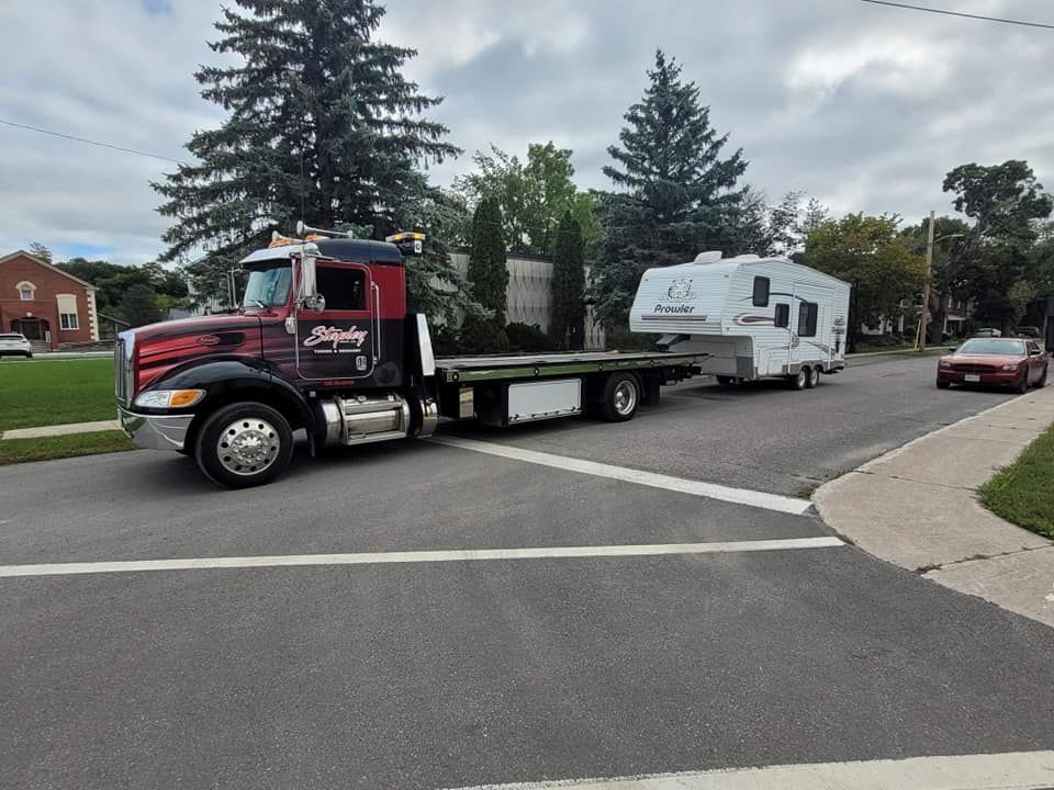 Tow truck towing a travel trailer on a residential street. Red truck with black details and a white trailer.