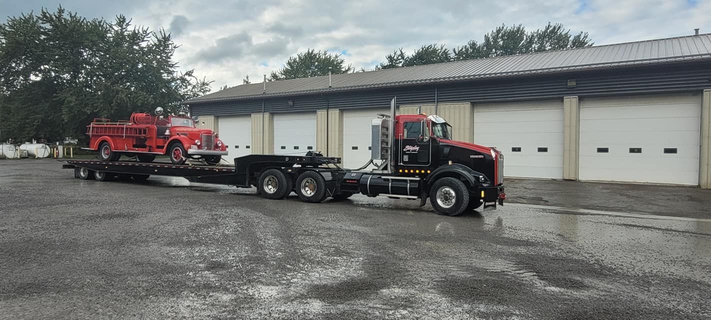 A red antique fire truck on a trailer towed by a black semi-truck parked in front of a building with garage doors.